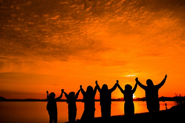 Silhouetted group raising arms against a vibrant sunset near a serene lake, capturing a sense of joy and unity.